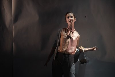 a woman in grey and pink stares out as a hand reaches through the wall behind her and touches her extended arm