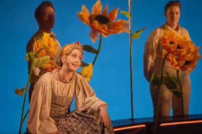 A woman is seen in sitting singing amongst large sunflowers with 2 onlookers out of focus in the background