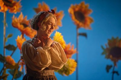 A woman is seen in distress standing hugging herself surrounded by large sunflowers