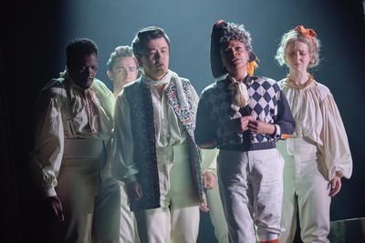 A group of 5 people are standing in a close group, all looking slightly downwards towards the floor, all grieving