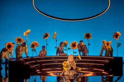 A striking blue background behind a semi circle of people watching the central action: a woman on the floor clasping a bunch of sunflowers