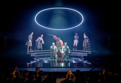 A semi circle of characters stand on a black metal platform with dark lighting, contrasting with the bright lighting of 2 people on their knees awaiting beheading by sword