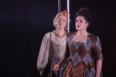 Two woman are seen wearing dresses. The woman in the foreground is singing distressed, and the other woman slightly out of focus, is looking at her, concerned