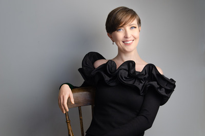 Sarah is seated on a wooden chair with an arm draped over from back to front, smiling widely