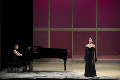 Sarah stands in a black shoulderless ball gown looking sad as she sings with a woman playing piano on stage at the Wexford Theatre